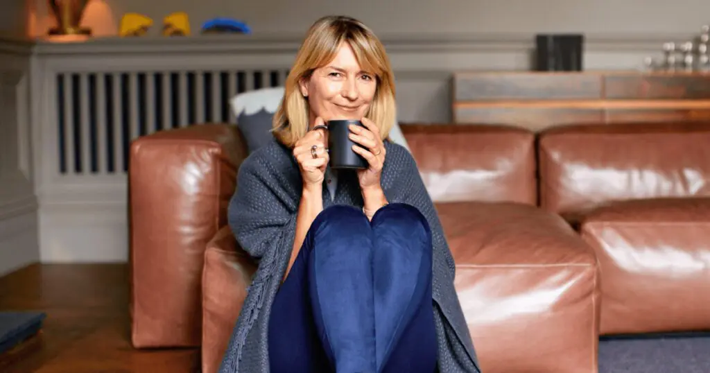 woman sitting in front of couch wrapped in blanket sipping warm drink