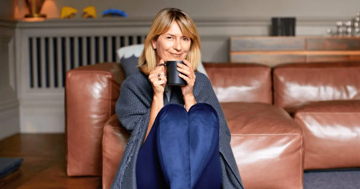 woman sitting in front of couch wrapped in blanket sipping warm drink